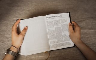 Twee handen houden kerkbijbel vast die ligt opengeslagen bij de eerste bladzij van de Nederlandse Geloofsbelijdenis.