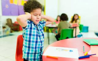 Jongen van een jaar of zeven in een klaslokaal houdt zijn handen tegen zijn oren; hij zit blijkbaar niet lekker in zijn vel.