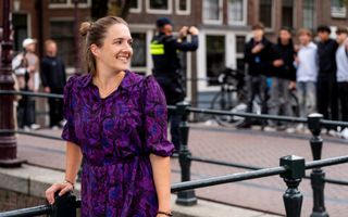Een jonge vrouw in paarse, gebloemde jurk, staat op een brug over een gracht. Op de achtergrond lopen mensen.