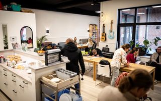 Een grote lichte ruimte met een tiental mensen dat zit of staat. Links een keukenblok, langs de muur drie banken, verschillende gitaren en een keyboard. In het midden een lange houten tafel.