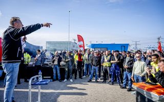 Een groep mannen in werkkleding luistert naar iemand met een microfoon in de hand, die hen toespreekt. Op de achtergrond staat een vlag van vakbond FNV.