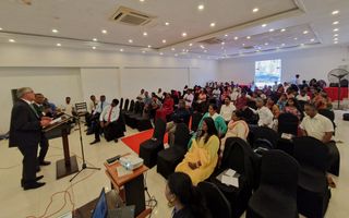 Aziatisch uitziende mensen luisteren in een conferentiezaal naar een Nederlandse spreker in pak.