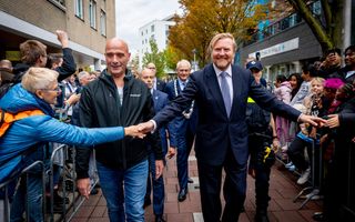 De koning loopt tussen dranghekken links en rechts door. Hij heeft beide handen uitgestoken en schudt zo aan beide zijden handen.
