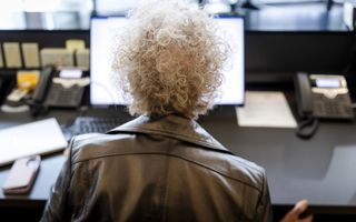 Een dame met grijs haar zit aan een bureau en kijkt naar een computerscherm. Je ziet haar op de rug.