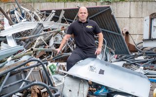 Kale man met tattoo op zijn arm, gekleed in zwart T-shirt en broek, staat tussen enorme rommel aan oud ijzer. 