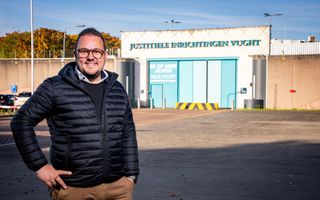Een man met een donkere jas aan en een bruine broek staat voor een pand waarop staat: justitiële inrichtingen Vught.
