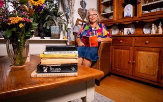 Oudere vrouw met kleurige blouse zit in een stoel met een boek in haar handen. Voor haar op de tafel ligt een stapel boeken. 