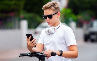 Jongen met blond haar en een zonnebril op kijkt op zijn mobiel. Hij houdt het stuur van zijn fiets met zijn linkerhand vast. Om zijn nek hangt een koptelefoon. 