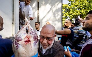 Een oudere man met een kort baardje heeft een groot stuk vlees, dat half uit een blauwe plastic zak steekt, in zijn armen. Op de achtergrond andere jonge mannen.
