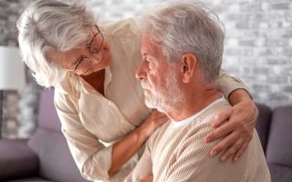 Een vrouw met grijs haar legt een arm over de schouder van een wat oudere man die verloren voor zich uit kijkt. 