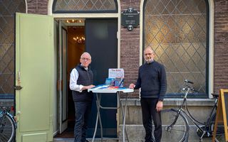Twee mannen staan voor de gevel van een kerkgebouw. Tussen hen in een statafel met evangelisatiemateriaal.