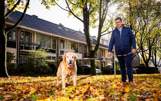 Een man met bril en een blauwe jas aan staat met een hond aan de riem staat buiten op een grasstrook met veel herfstbladeren. Op de achtergrond staan rijtjeshuizen.