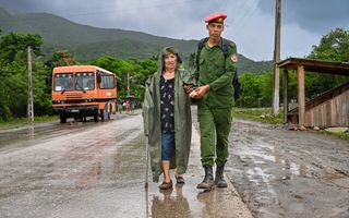 Een oudere vrouw en een militair lopen op een asfaltweg. Links een bus. Op de achtergrond bomen en heuvels.