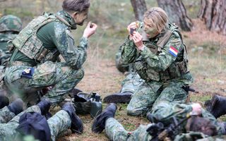 Koningin Máxima zit op haar knieën in een bosachtige omgeving in militair tenue. Met een klein sponsje smeert ze groene verf op haar gezicht, terwijl ze in een zakspiegeltje kijkt. Om haar heen anderen in militair uniform.