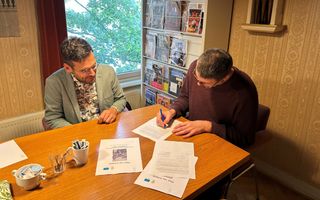 Twee mannen zitten aan een tafel en zijn bezig met het ondertekenen van een overeenkomst.