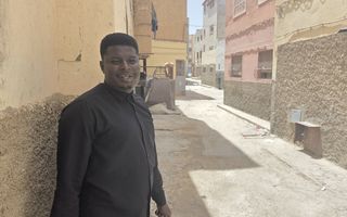 Man in zwarte kleding staat in een smalle straat tussen pastelgekleurde huizen.