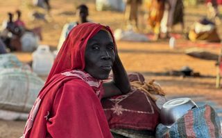 Donkere vrouw, met een rode doek over haar hoofd, leunt met een arm op wat samengeknoopte bezittingen.