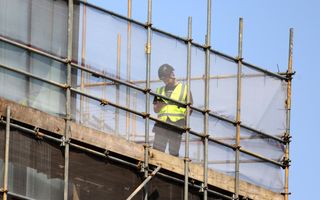 Een man draagt een veiligheidsvest en staat op een steiger. De steiger bestaat uit stalen delen, verbonden met een houten balk.