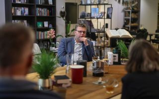 Man met overhemd, blauw colbert en bril zit aan een tafel tegenover een man en een vrouw. Op de achtergrond een boekenkast en een grote, opengeslagen Bijbel op een standaard.