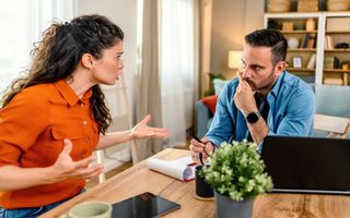 Gefrustreerd stel dat thuis ruziemaakt. Vrouw links in feloranje blouse probeert al gebarend iets duidelijk te maken aan man met lichtblauwe blouse achter laptop en papieren.