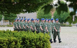 Jonge mannen en vrouwen marcheren op een plein omringd door bomen en een heg in camouflagegekleurd legeruniform. Rechterarmen en linkerbenen staan voor. Alle rekruten dragen een blauwe baret.