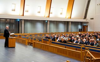 Een kerkzaal met mensen; links dominee Schot achter een katheder.
