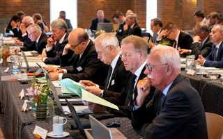 Mannen in donkere pakken achter lange vergadertafels. Voor hen koffiekopjes, glazen water, papieren en laptops.