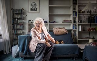 Oudere vrouw met licht-roze jasje en grijze krullen zit op een bank waar een blauw-zwart geblokt kleed op ligt. Naast haar op de bank ligt een hondje. Op de achtergrond staan wat kasten die deels leeg zijn.