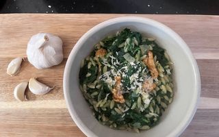 Schaaltje met een groen gerecht, bestrooid met kaassnippers. Het schaaltje staat op een houten plank waar ook een bol knoflook en knoflookteentjes op liggen.