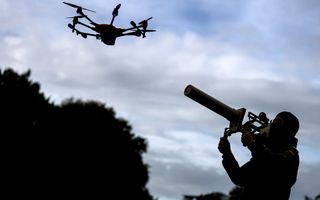 Links een in het donker geklede man met een bivakmuts op houdt een langwerpig wapen op zijn schouder en richt dat op een drone die schuin voor hem vliegt.