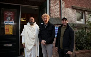 Drie mannen staan naast elkaar voor de ingang van een gebouw. De man links heeft een baard en draagt witte kleding. 