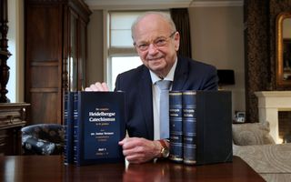 Een oudere, kalende man, zit achter een tafel. Voor hem staan vier dikke boeken.