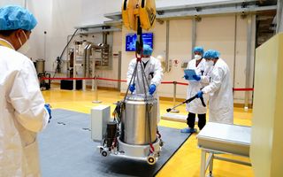 Mensen met witte labjassen aan, met mondkapjes voor en blauwe haarnetten op hun hoofd verplaatsen een metalen cilinder met een ingebouwde kraan. 