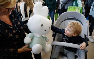 Een kindje in een buggy grijpt naar een grote pop van Nijntje die een vrouw vasthoudt.