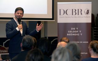 Man in blauw pak en witte blouse spreekt in microfoon naast banner met letter DCBR erop.