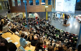 Een grote studiezaal, met een tribune vol mensen en voorin een podium en projectiescherm.