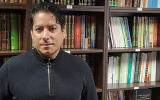 Man met zwart haar, trui, voor een boekenkast.