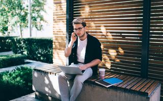 Jonge man met donker haar en een bril zit met een laptop op schoot op een bankje in een tuin. Hij heeft een telefoon aan zijn oor. Achter hem is een houten scherm. Op het bankje staat een beker koffie.