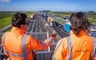 Twee mannen in een oranje jas staan op een viaduct boven een brede weg waarop vrachtwagens en auto’s te zien zijn.