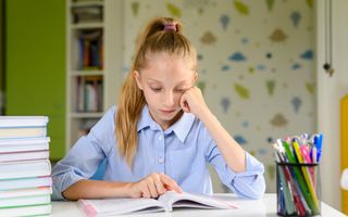 Meisje met een paardenstaart zit in een boek te lezen. Ze houdt haar wijsvinger bij de plek waar ze is. Haar linkerhand houdt ze gebald tegen haar wang.