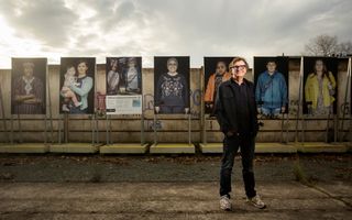 Man met zwarte kleding en bril staat voor een serie portretfoto’s die op standaards voor een betonnen muur staan.