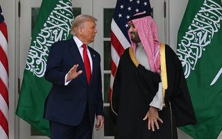 Twee mannen, de een in pak en de ander in zwart gewaad met rood geruite hoofddoek, staan voor een wit gebouw. Achter hen zijn Amerikaanse en Saudische vlaggen te zien