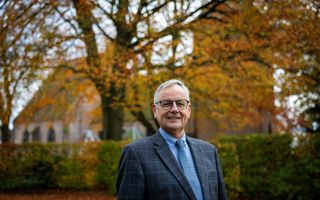 Dominee Wassenaar, een man met grijs haar, bril op, gekleed in het blauw, met op de achtergrond bomen in herfstkleuren en een oude kerk.