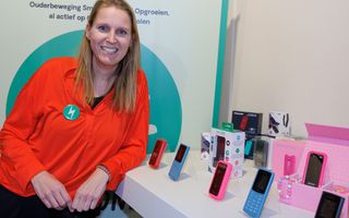 Blonde vrouw in oranje shirt bij een tafel waarop allerlei gekleurde eenvoudige telefoons staan uitgestald.