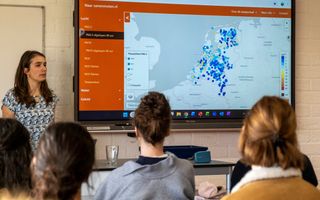 Een vrouw staat voor een groep leerlingen. Naast haar een smartboard met een kaartje van Nederland vol blauwe en gele stippen. 