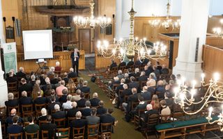 Kerkzaal met houten stoelen gevuld met mensen, spreker in pak staat voorin en kroonluchters hangen aan het plafond.
