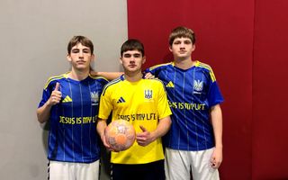 Drie jongeren in sportkleding staan naast elkaar. De jongeren hebben donkerblond of bruin haar en dragen geel met blauwe shirts. Daarop staat de tekst: Jesus is my life. De middelste jongen houdt een voetbal vast.