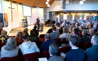 Een zaal vol mensen, schuin van achteren gezien. Voorin staat een man in donker pak achter een katheder. Aan de wand achter hem hangt een groot kruis.