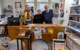 Drie mensen achter een houten toonbank. Overal in de winkel boeken, kaarten en ander geschenkmateriaal.
