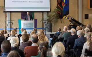 Zaal vol mensen op de rug gezien. Voorin staat een man in een donker pak achter een katheder.  